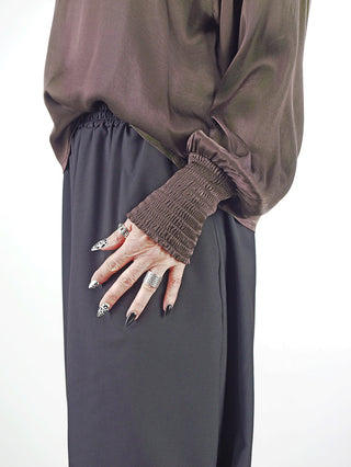 Person trägt das OFF#DLY LS Shirt Valentina braun mit fließendem Stoff und weiten Ärmeln mit gerafften Bündchen, kombiniert mit hoch taillierten schwarzen Hosen und auffälligen silbernen Ringen.