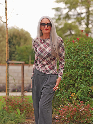 Ältere Frau im Garten trägt das OFF#DLY LS light m. Cashmere Karo bordeaux, ein langärmeliges, zartes Oberteil mit mutedem Karo-Muster und femininer Silhouette, kombiniert mit grauen Hosen.