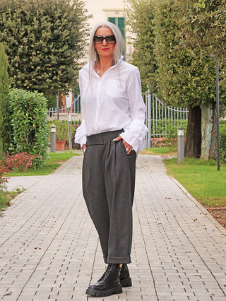 Eine Frau trägt die OFF#DLY Bluse Viola white mit rüschigen Ärmeln, kombiniert mit hochgeschnittenen, weiten Hosen und robusten Stiefeln, in einem gepflegten Gartenweg.