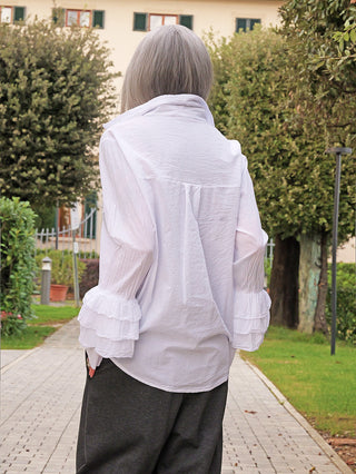 Rückansicht einer Person, die die OFF#DLY Bluse Viola white trägt. Die Bluse hat lockere, mehrlagige Volant-Ärmel und einen luftigen Schnitt, kombiniert mit dunklen, weiten Hosen.