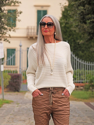 Ältere Frau im OFF#DLY Pulli Cara offwhite, einem cremefarbenen, gerippten Pullover, steht mit Händen in den Taschen auf einem Weg. Der Pullover ergänzt stilvoll ihre braunen Hosen und Perlenkette.