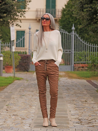 Ältere Frau trägt OFF#DLY Joggpant Antonia camel mit Knitter-Look, hoher Taille und Bindeband, kombiniert mit lockerer Elfenbein-Pullover, Sonnenbrille und Stiefeletten vor einer Villa und Tor.