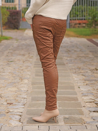 Die OFF#DLY Joggpant Antonia camel zeigt sich im modernen Knitter-Look, schmaler Passform und elastischem Komfortbund, getragen mit cremefarbenen Stiefeletten auf einem gepflasterten Weg.