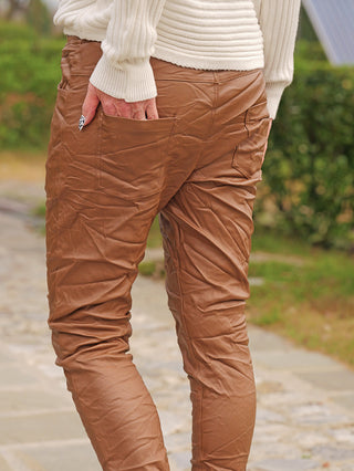 Eine Person trägt OFF#DLY Joggpant Antonia camel mit Knitterlook, schmalem Bein und Gesäßtasche. Ihre Hand steckt in der rechten Tasche, ideal für moderne Alltags-Outfits.