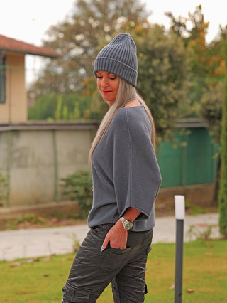 Frau trägt OFF#DLY Pulli Ellen Asphalt mit lockerer Passform und Fledermausärmeln, ergänzt durch eine Rippstrickmütze und Cargo-Hosen, in einem Garten stehend.