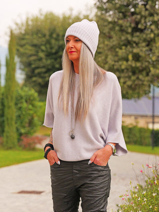 Person im Freien trägt eine OFF#DLY Mütze Anna eisgrau, kombiniert mit dunkel strukturierten Hosen, einem hellen Pullover. Accessoires: lange Halskette, Armbanduhr, und Perlenarmbänder.
