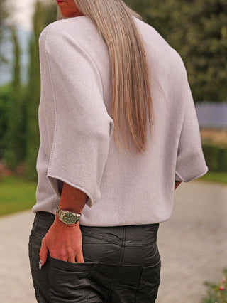 Person von hinten in OFF#DLY Pulli Ellen eisgrau mit lockerer Passform und Fledermausärmeln, kombiniert mit schwarzen Lederhosen und einer dunklen Armbanduhr, steht auf einem Weg im Grünen.