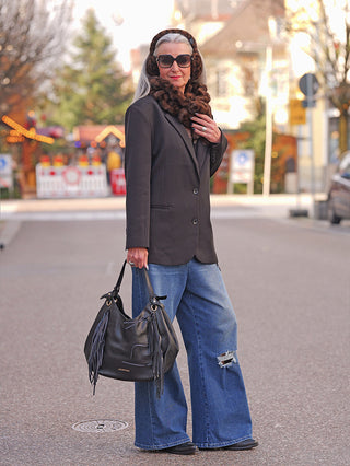Ältere Frau in urbanem Setting trägt OFF#DLY Ohrwärmer black/leo in Kunstpelz über Kopf und Hals, kombiniert mit dunklem Blazer, Jeans und Sonnenbrille, hält schwarze Fransenhandtasche.