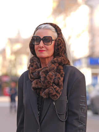 Frau mit silbernem Haar trägt den OFF#DLY Fur Schal black/leo aus weichem Kunstfell, kombiniert mit passenden Ohrenschützern, über einem dunklen Blazer mit Kettendetail.