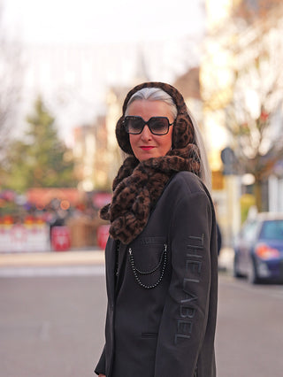 Frau trägt OFF#DLY Ohrwärmer black/leo, weiche, schwarze Leopard-Print-Fake-Fur-Ohrwärmer, kombiniert mit passendem Schal, oversized Sonnenbrille und rotem Lippenstift, während sie auf einer städtischen Straße steht.