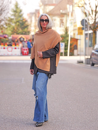 Ältere Frau trägt den OFF#DLY Poncho Lilly camel, weich drapiert über schwarzer Lederjacke, kombiniert mit Jeans und Stiefeletten, was den eleganten, fließenden Stil des Ponchos betont.