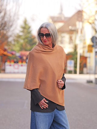 Eine Frau trägt den OFF#DLY Poncho Lilly camel über einer schwarzen Jacke. Der Poncho ist leicht und asymmetrisch, ideal für stilvolle, elegante Looks.
