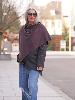 Ältere Frau trägt den OFF#DLY Poncho Lilly schoko, asymmetrisch drapiert über schwarzem Blazer, kombiniert mit lässigen Jeans. Ideal für stilvolle, warme Übergangslooks in der Stadt.