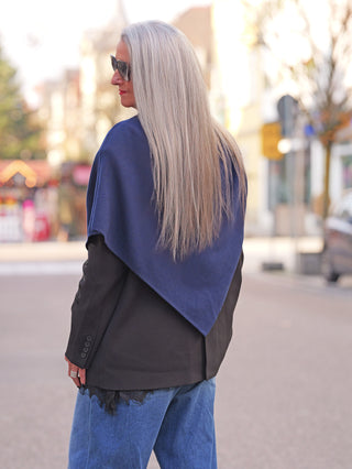 Eine Person trägt den OFF#DLY Poncho Lilly marine über einem schwarzen Blazer, kombiniert mit weiten Jeans, und spaziert in einer städtischen Umgebung.