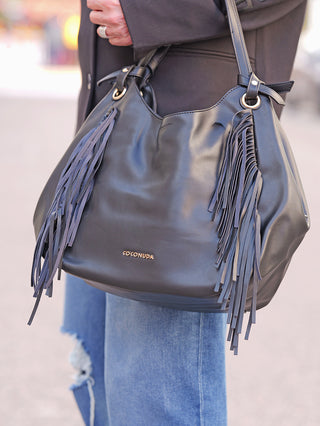 Nahaufnahme der Cocondua Fransen Bag black, eine schwarze Lederhandtasche mit langen Fransen an den Seiten. Sie trägt ein kleines goldfarbenes COCONUDA-Logo auf der Vorderseite.