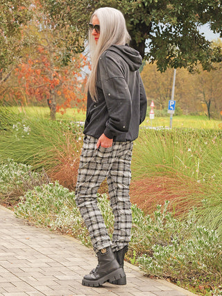 Person mit silbergrauen Haaren und Sonnenbrille trägt den OFF#DLY Hoody Pia black washed, einen lässigen, schwarzen Kapuzenpullover, kombiniert mit schwarz-weißen karierten Hosen und klobigen Boots auf einem herbstlichen Weg.