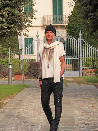 Person trägt OFF#DLY Pulli m. Schal Carla offwhite, locker sitzender Strickpulli mit passendem Schal, ergänzt durch schwarze Hose und Stiefel, vor einem Haus mit Garten.