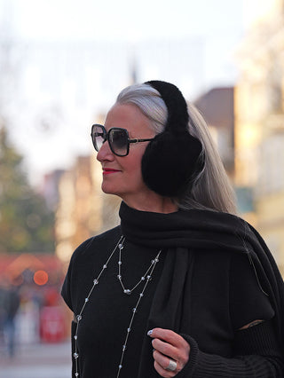 Ältere Frau mit langen silbernen Haaren trägt OFF#DLY Ohrwärmer black aus Kunstfell, kombiniert mit schwarzen Sonnenbrillen, schwarzem Outfit und rotem Lippenstift, in städtischer Umgebung.
