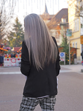 Person von hinten mit langen, silbrig-grauen Haaren trägt den OFF#DLY Blazer Fiora black, schmal geschnitten mit Broschen am Kragen. Hintergrund zeigt unscharfen, festlichen Marktplatz.