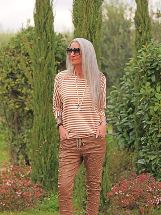 Frau trägt das OFF#DLY Ringelshirt Cincia camel/beige, locker geschnitten mit beige-weißen Streifen, leicht oversized. Kombiniert mit brauner Hose, betont es Komfort und Stil in einem eleganten Parkambiente.