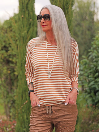 Ältere Frau im Garten trägt das beige-weiße Ringel Donna Shirt von OFF#DLY mit lässigem Schnitt und langen Ärmeln, kombiniert mit braunen Hosen und einer langen Halskette.