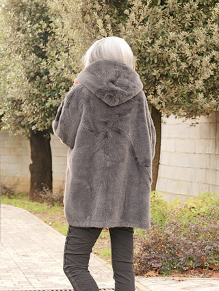 Eine Person trägt die OFF#DLY FakeFur Jacke Rom asphalt, eine graue, flauschige Kapuzenjacke, während sie auf einem gepflasterten Weg spaziert.