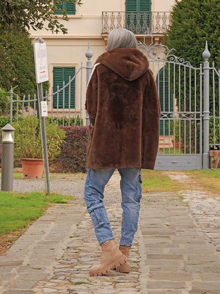 Eine Person trägt die OFF#DLY FakeFur Jacke Rom braun, eine schokoladenbraune, knielange Kunstpelzjacke mit Kapuze, während sie auf einem Gartenweg in Richtung eines kunstvollen Tores läuft.