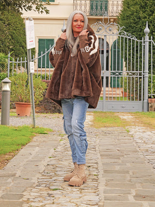 Eine Frau trägt die OFF#DLY FakeFur Jacke Rom braun, oversized mit Kapuze, über hellblauen Cargo-Jeans und klobigen Stiefeln, während sie einen steinernen Weg entlanggeht.