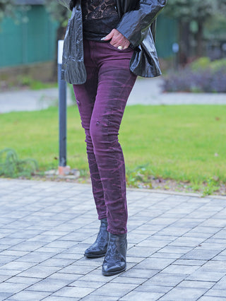 Person trägt auf einem gepflasterten Weg die OFF#DLY Denim Eva bordeaux, eine figurbetonte, tief auberginefarbene Hose mit leichten Distressed-Details, kombiniert mit schwarzen Lederstiefeletten und offenem, glänzendem Mantel.