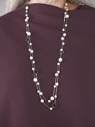 Silberne OFF#DLY Kette Riga mit Kunstperlen und schwarzen Kleeblatt-Charms, auf einem pflaumenfarbenen Oberteil präsentiert. Die Kette zeigt einen abwechselnden Perlen- und Kleeblatt-Motiv-Muster.