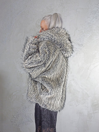 Person in Profilansicht trägt eine OFF#DLY Fake Fur Jacke creme/black mit hochgezogenem Kragen, silber-grauem Pferdeschwanz und einem dunklen, gemusterten Kleid darunter, vor einer leicht strukturierten Wand.