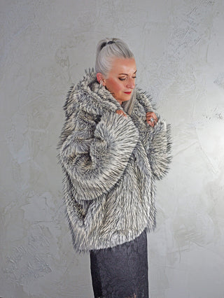 Eine Frau trägt die OFF#DLY Fake Fur Jacke creme/black mit voluminösem Kragen. Sie hält den weichen Stoff um ihre Schultern, während ein dunkler Spitzenrock darunter hervorschaut.