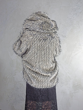 Eine Person trägt die OFF#DLY Fake Fur Jacke creme/black, eine dicke, kapuzenbesetzte Kunstfelljacke in Grautönen. Darunter ist ein langer, dunkler Spitzenrock oder -kleid sichtbar.