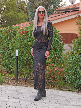 Eine Person trägt das OFF#DLY Rock Resi black, ein langes, figurbetontes schwarzes Spitzekleid mit floralen Mustern, kombiniert mit schwarzen Stiefeletten und silberner Halskette, in einem Garten.