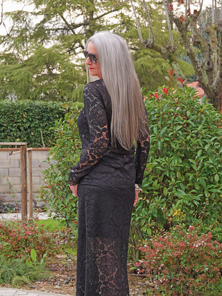 Person im Garten trägt eine lange schwarze Spitzenkleid, passend zum OFF#DLY Rock Resi black, mit floralen Mustern und transparenten Partien, die moderne Eleganz mit femininer Spitze kombinieren.