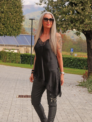 Person in Park trägt OFF#DLY Top Spitze Veronika black, ein asymmetrisches, fließendes schwarzes Tunika mit Spitzensaum, kombiniert mit figurbetonten schwarzen Hosen mit Zipper-Details.