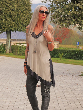 Frau mit langem, grauem Haar trägt das OFF#DLY Top Spitze Veronika gold/black, ein fließendes, ärmelloses Tunika-Top mit asymmetrischem Saum und Spitzenakzenten, kombiniert mit schwarzen Hosen.