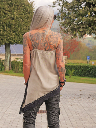 Person von hinten in OFF#DLY Top Spitze Veronika gold/black, locker geschnitten, mit asymmetrischem Spitzensaum, kombiniert mit schwarzen Hosen. Sichtbare Tätowierungen und lange graue Haare.