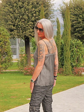 Frau mit langen, silbernen Haaren trägt das OFF#DLY Top Spitze Allegra asphalt, ein ärmelloses, weiches Top mit edler Spitzenbordüre, ideal für feminine, moderne Layering-Outfits in einem Park.
