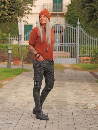 Person trägt OFF#DLY Pulli Tina zimt, ein rustfarbenes, kurzärmeliges Strickoberteil. Kombiniert mit schwarzen Cargo-Hosen und Boots, vor einer Villa mit schmiedeeisernem Tor und gepflegtem Garten stehend.