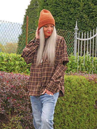 Person mit grauem Haar trägt OFF#DLY Mütze Anna zimt, eine rost-orangefarbene, gerippte Beanie, kombiniert mit braunem, kariertem Hemd und zerrissenen Jeans, stehend vor Hecke im Garten.