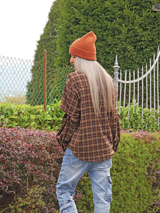 Person von hinten, trägt die OFF#DLY Bluse Hilde braun, ein oversized kariertes Hemd, in einem Garten stehend. Kombiniert mit orangefarbener Beanie und blauen Cargo-Jeans für einen lässigen Look.