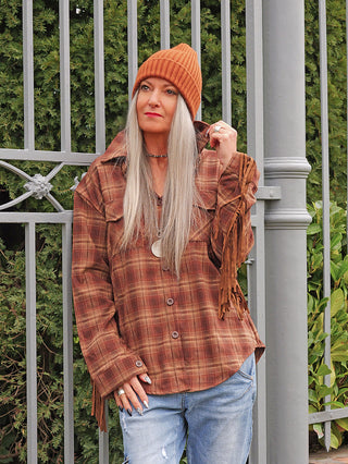 Erwachsene in orangefarbener Mütze trägt die OFF#DLY Bluse Rodeo braun mit Fransen, kombiniert mit hellen Jeans, lehnt lässig an einem Zaun, zeigt modernen Westernstil.