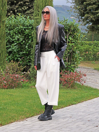 Frau trägt OFF#DLY Hose Moniga weiß, kombiniert mit einem schwarzen Blazer und Spitzen-Top, auf einem Gartenweg. Die Hose ist weit geschnitten mit asymmetrischem Bund und dekorativen Knöpfen.