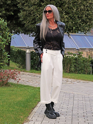 Person trägt OFF#DLY Hose Moniga white, hoch-tailliert mit asymmetrischem Bund und Knopfdetails, kombiniert mit schwarzem Spitzen-Top und Lederjacke, vor Gartenhintergrund mit modernen Elementen.