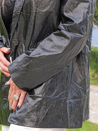 Nahaufnahme einer Person im OFF#DLY Fake Leather Blazer Gina black mit glänzender Crinkle-Optik, gefalteten Händen mit gemusterten Nägeln und silbernem Ring, kombiniert mit hellen Hosen.