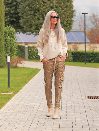 Ältere Frau trägt den OFF#DLY Hoody (Strick) Daria beige, mit dezenter Logo-Stickerei und Kapuze, während sie auf einem gepflasterten Weg in einem gepflegten Garten spaziert.