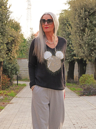 Frau mit langem silbergrauem Haar trägt den OFF#DLY Pulli Kira black mit Mickey-Mouse-Applikation und goldenen Details, kombiniert mit grauen Hosen und goldenen Accessoires, während sie einen Parkweg entlanggeht.