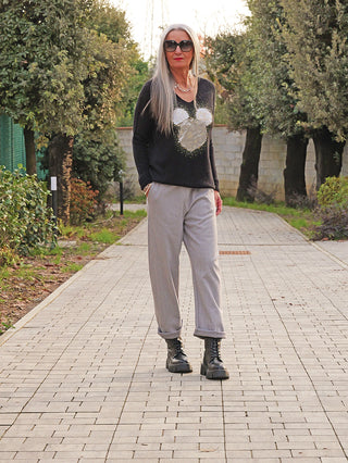 Frau in OFF#DLY Pulli Kira black mit Mausmotiv, goldenen Details und Strass, locker fallend. Kombiniert mit grauen Hosen und schwarzen Stiefeln auf baumbewachsenem Weg spazierend.