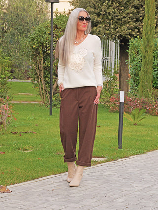 Frau trägt den OFF#DLY Pulli Kira white mit großem goldverziertem Motiv. Der Pullover aus weicher Baby-Alpaka-Mischung kombiniert Eleganz mit Komfort auf einem Gartenweg spazierend.